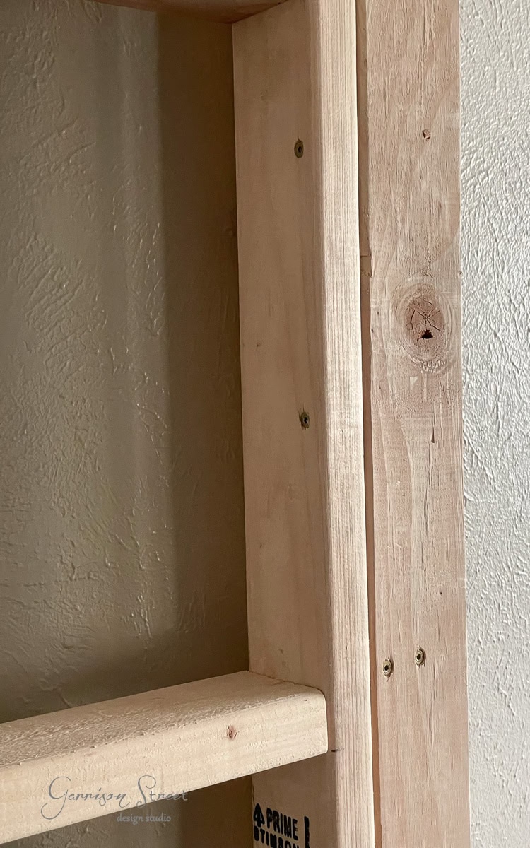 Close-up of a wooden bookshelf corner, focusing on the construction details. The unfinished, natural wood, ideal for a DIY electric fireplace and TV wall project, shows visible screws and knots. A subtle texture is seen on the adjacent beige wall.