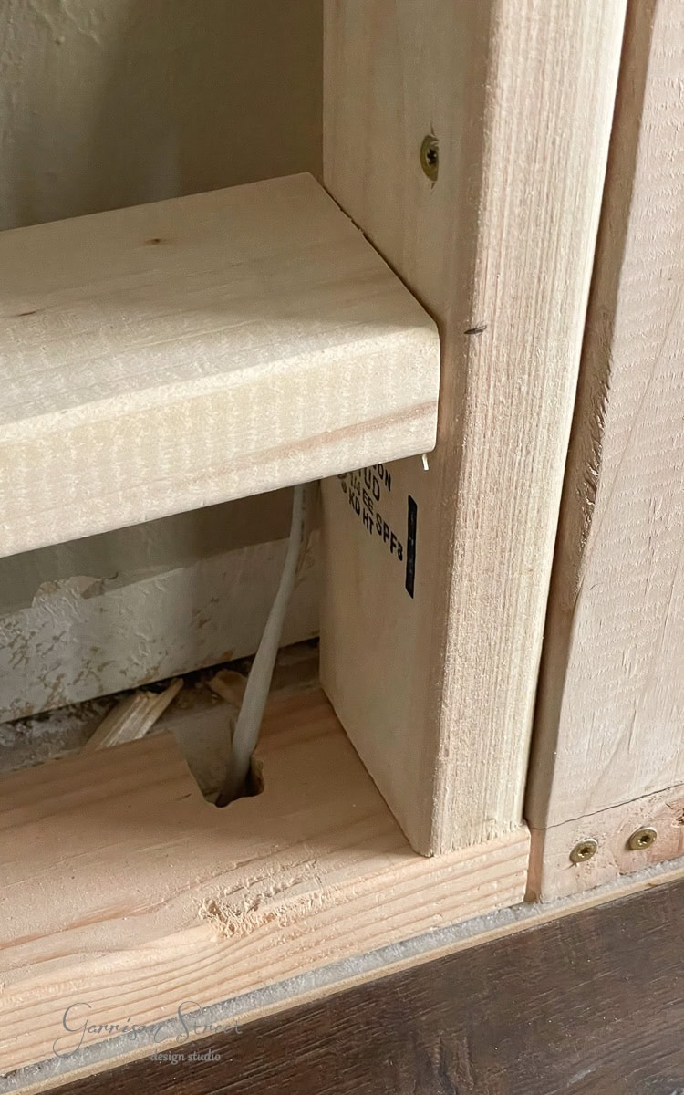 A partially constructed wooden frame forms the base of a DIY electric fireplace and TV wall, with planks joined together seamlessly. A white cable threads through a hole in the base, while visible screws secure the structure. Some text is delicately printed on the wood surface.