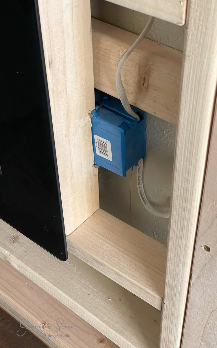 A wooden frame structure with a blue electrical box and exposed wiring is shown, hinting at the early stages of a DIY electric fireplace and TV wall project. A piece of black material is partially visible on the left side, adding a touch of mystery to the evolving setup.