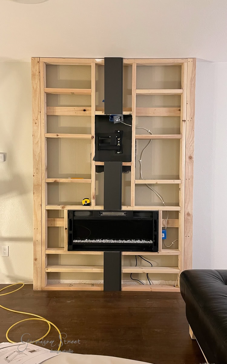 A partially constructed DIY electric fireplace and TV wall showcases a built-in electric fireplace and a mounted TV bracket. Exposed wiring is visible, surrounded by a section of a brown sofa and a yellow extension cord on the floor.