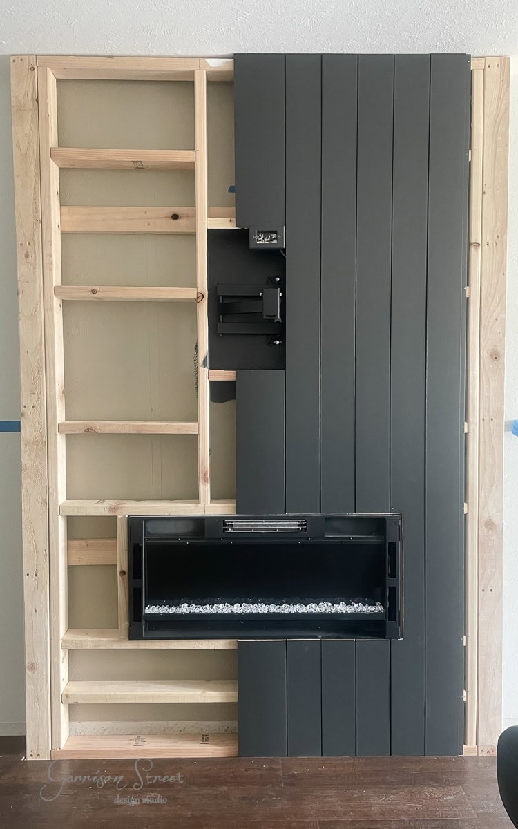 A DIY electric fireplace and TV wall, this partially constructed installation showcases unfinished wooden framing, a mounted TV bracket, and a recessed electric fireplace adorned with decorative stones. One section of the wall is elegantly covered with vertical dark gray panels.