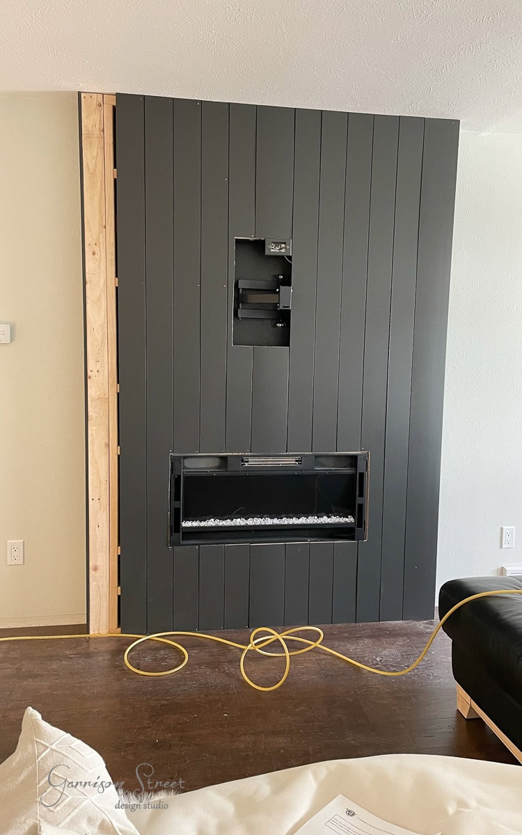 A partially finished, modern DIY wall installation features a black panel designed for an electric fireplace and TV mount. An exposed wooden frame can be seen on the left, while a yellow extension cord lies on the floor.