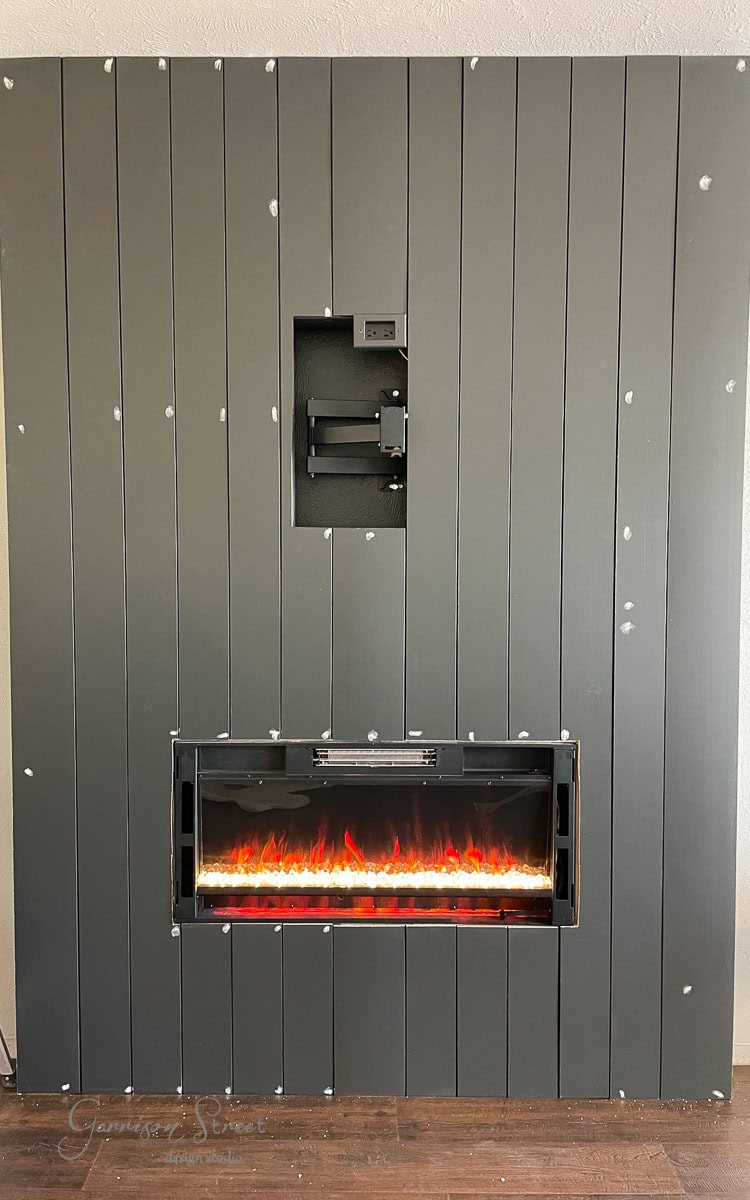 A modern DIY wall combines vertical dark gray panels with a built-in electric fireplace, showcasing glowing orange flames below. Above the fireplace lies a ready-to-use mount for your TV or monitor.