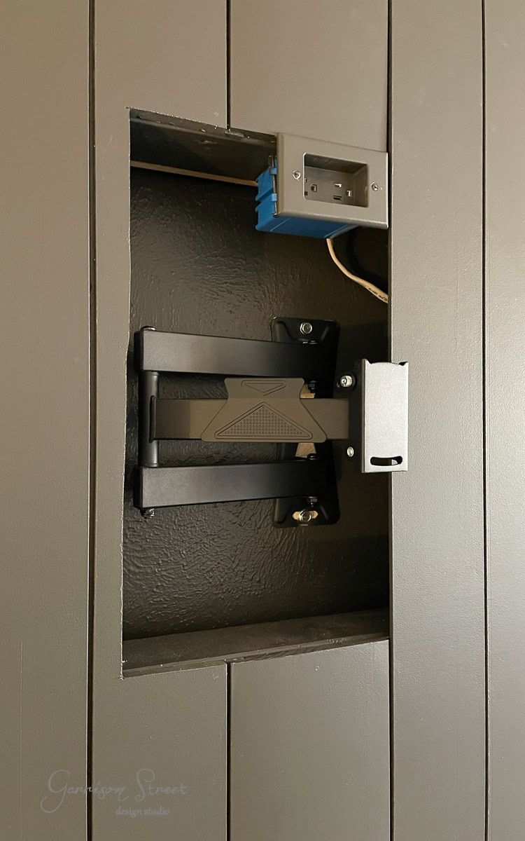 A black TV wall mount is installed inside a recessed space in a dark-colored wall, likely part of a DIY electric fireplace and TV wall project. Above the mount, an exposed electrical box with wires suggests the setup is a work in progress for television installation.
