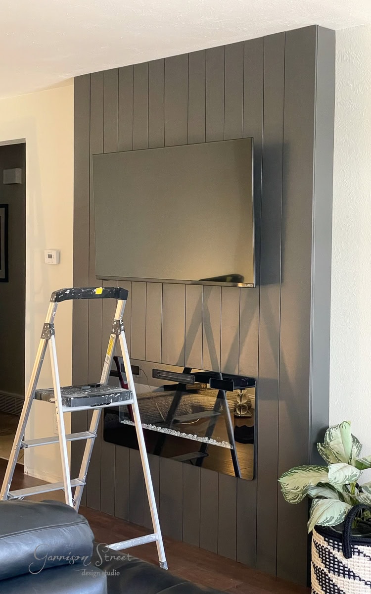 A modern living room showcases a DIY electric fireplace and TV wall. The gray vertical panel accent hosts a mounted flat-screen, while the sleek black fireplace is seamlessly integrated below. A silver step ladder stands nearby, adding character beside the floor-set plant.