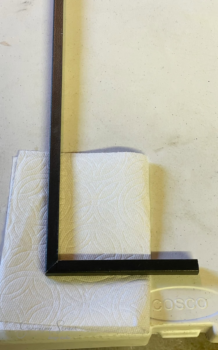 A black square metal frame in an L-shape rests on a piece of white textured paper towel atop a light gray surface, akin to the sleek setup of a DIY electric fireplace and TV wall. The scene casts subtle shadows, with a partial circular indentation visible on the surface.
