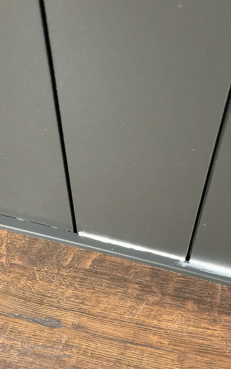 Close-up of a dark-colored, vertical panel wall meeting a wooden floor with a rich brown finish. Perfect for a DIY electric fireplace and TV wall, the panels are separated by thin, dark lines, while the floor exhibits a textured grain.