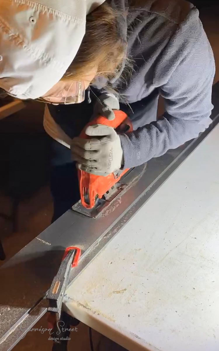 A person in protective gear carefully uses a jigsaw to cut metal clamped to a workbench, perhaps working on their next DIY electric fireplace and TV wall. They remain focused, donning gloves, a hood, and safety goggles for safety.