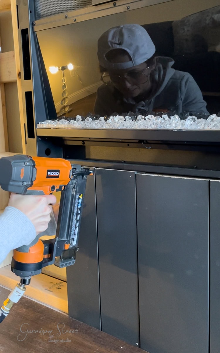 A person is using a nail gun on a wall panel under a television, crafting a sleek DIY electric fireplace and TV wall. They're wearing a cap and glasses, their reflection visible on the screen. Decorative rocks enhance the modern aesthetic of the space.