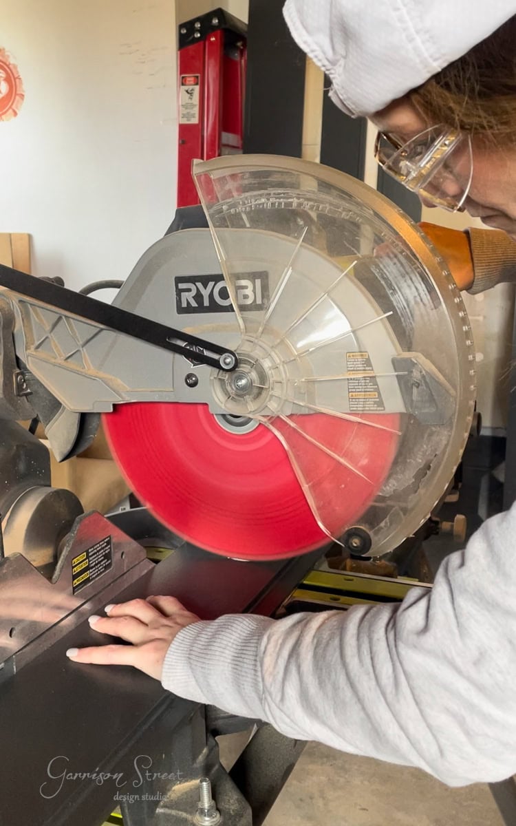 In a workshop setting, a person wearing safety glasses skillfully operates a Ryobi miter saw with a bright red blade. Their hand is steady on the cutting platform, crafting pieces for an upcoming DIY electric fireplace and TV wall project.