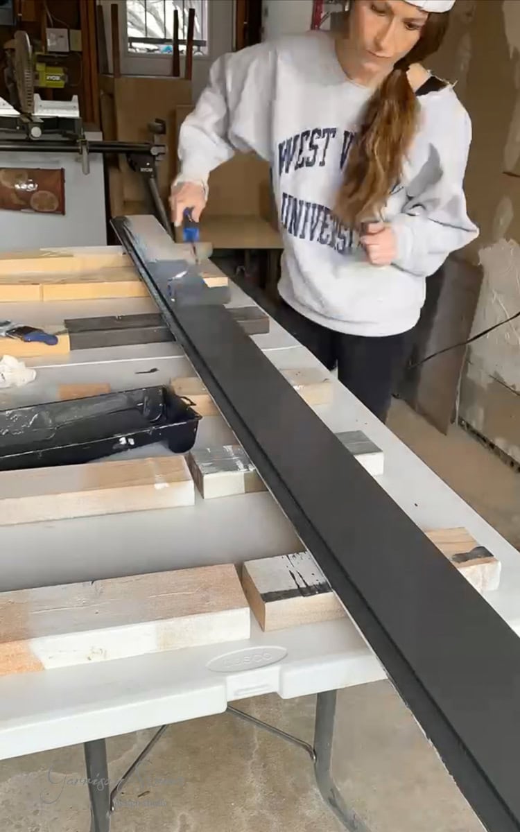 A person wearing a "West Virginia University" sweatshirt is in the workshop, painting a long wooden plank black for their DIY electric fireplace and TV wall project. The plank is supported by multiple wooden blocks on a table, showcasing their dedication to creating the perfect centerpiece.