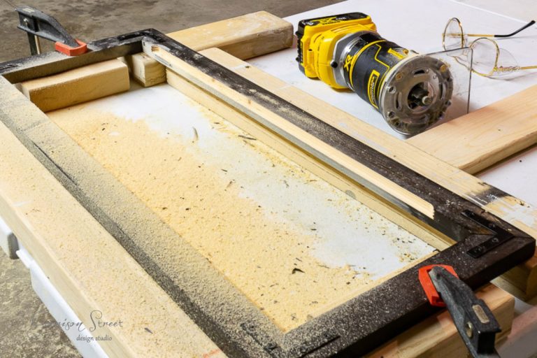 A woodworking setup showcases a yellow power router, among the essential tools for any DIY enthusiast, with a frame clamped to a table and sawdust scattered around. Safety glasses hang nearby, completing the scene in this bustling workshop.