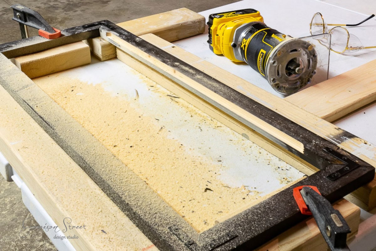 A woodworking setup showcases a yellow power router, among the essential tools for any DIY enthusiast, with a frame clamped to a table and sawdust scattered around. Safety glasses hang nearby, completing the scene in this bustling workshop.
