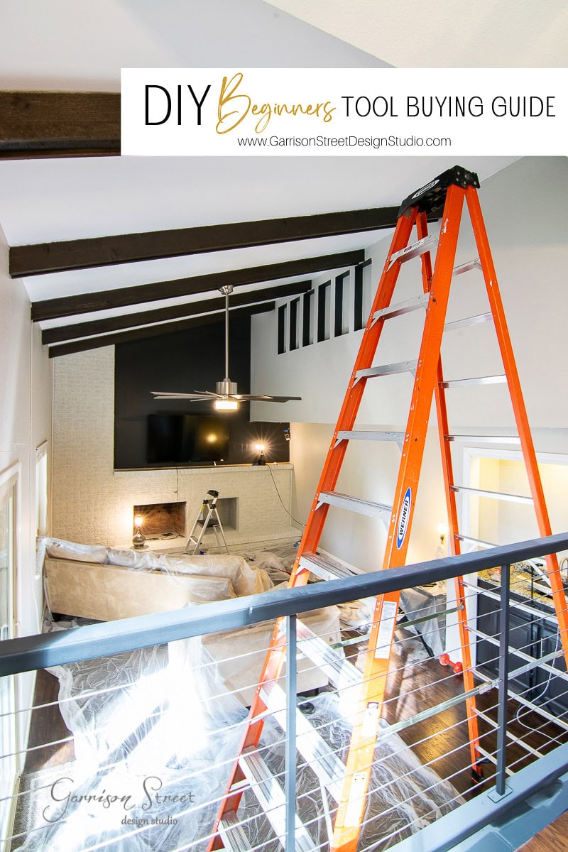 A modern living room with high ceilings is under renovation. An orange ladder stands near a couch covered with a drop cloth. A TV is mounted above the fireplace.