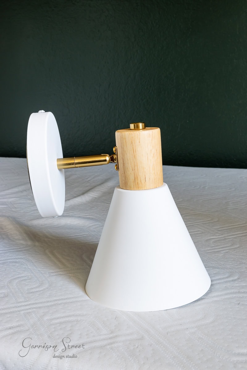 Displayed against a textured white surface, this wall sconce features a white conical shade, wooden top, and brass arm. Set against a dark green wall.