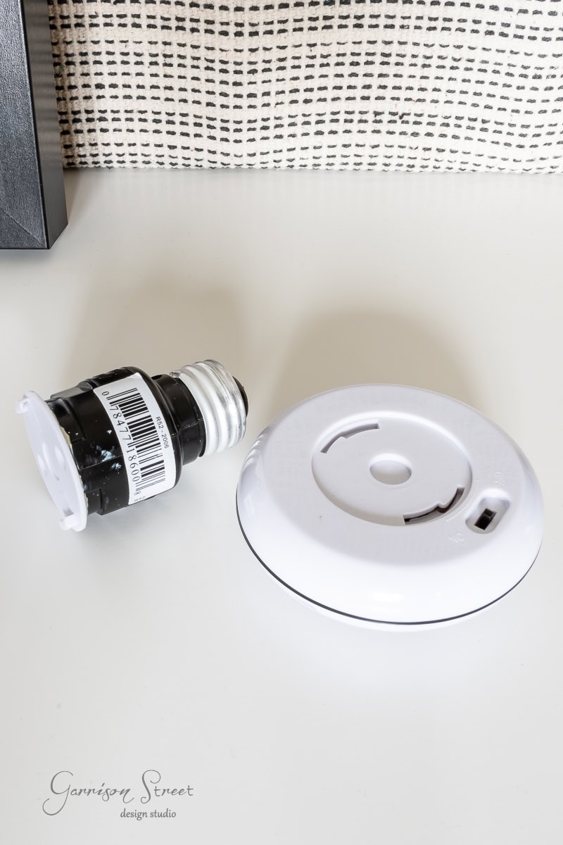 A detached smoke detector with its cover and base rests on a white surface. Behind it is a rolled-up piece of black tape with a barcode, while patterned fabric adds texture to the scene.