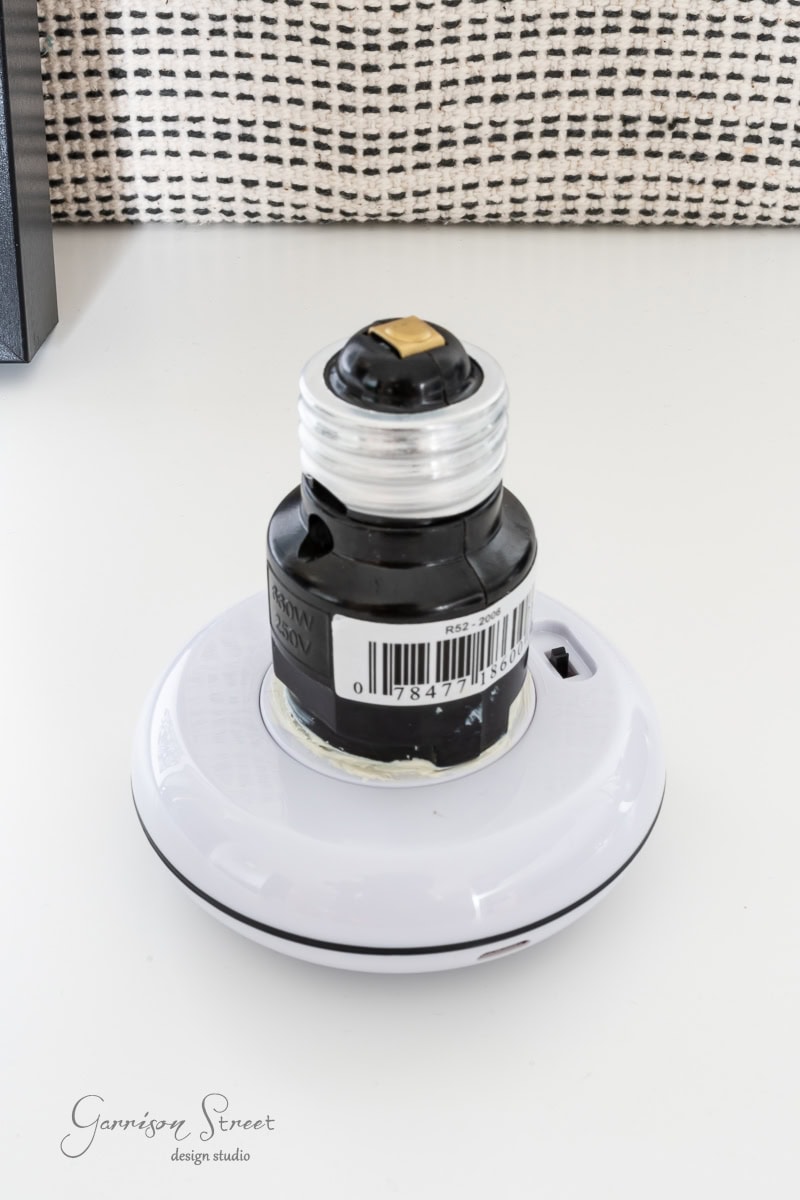 A white smart light bulb adapter sits elegantly on a table, framed by a black and white patterned wall. The adapter features a top socket and an easy-access on/off switch.