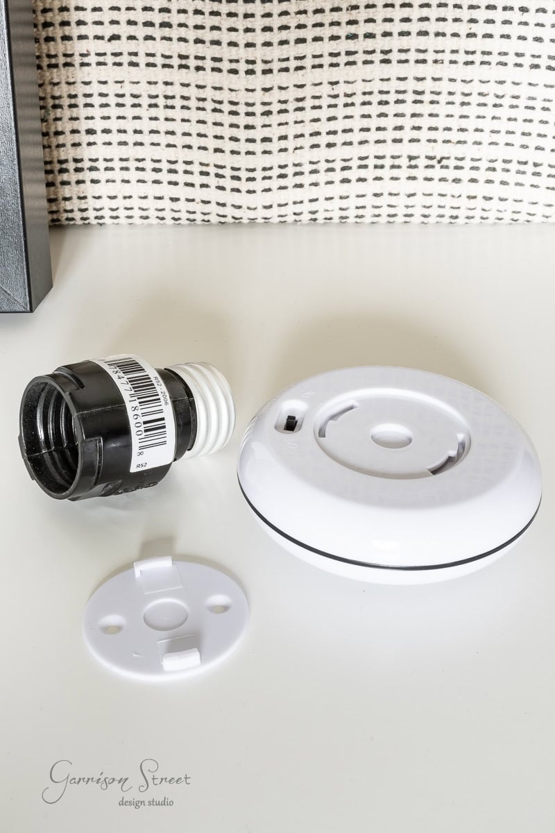 A disassembled light socket and adaptor with a black cylindrical component, a white circular base, and a small round mounting plate are arranged on a white surface. In the background, textured fabric adds texture to the scene.