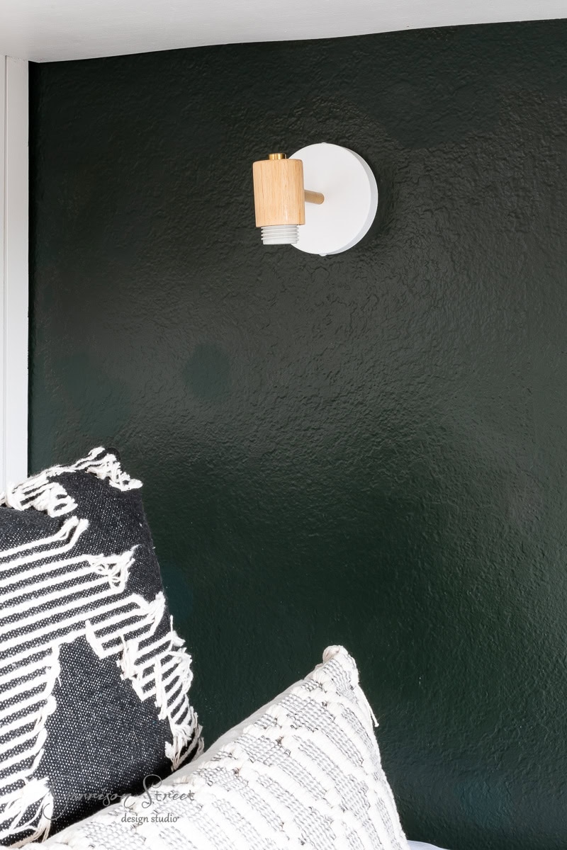 A modern DIY wireless wall sconce with a wooden accent complements the dark green wall. Below, a cushioned seating area features two patterned pillows, one in black and white and the other in white with black designs.