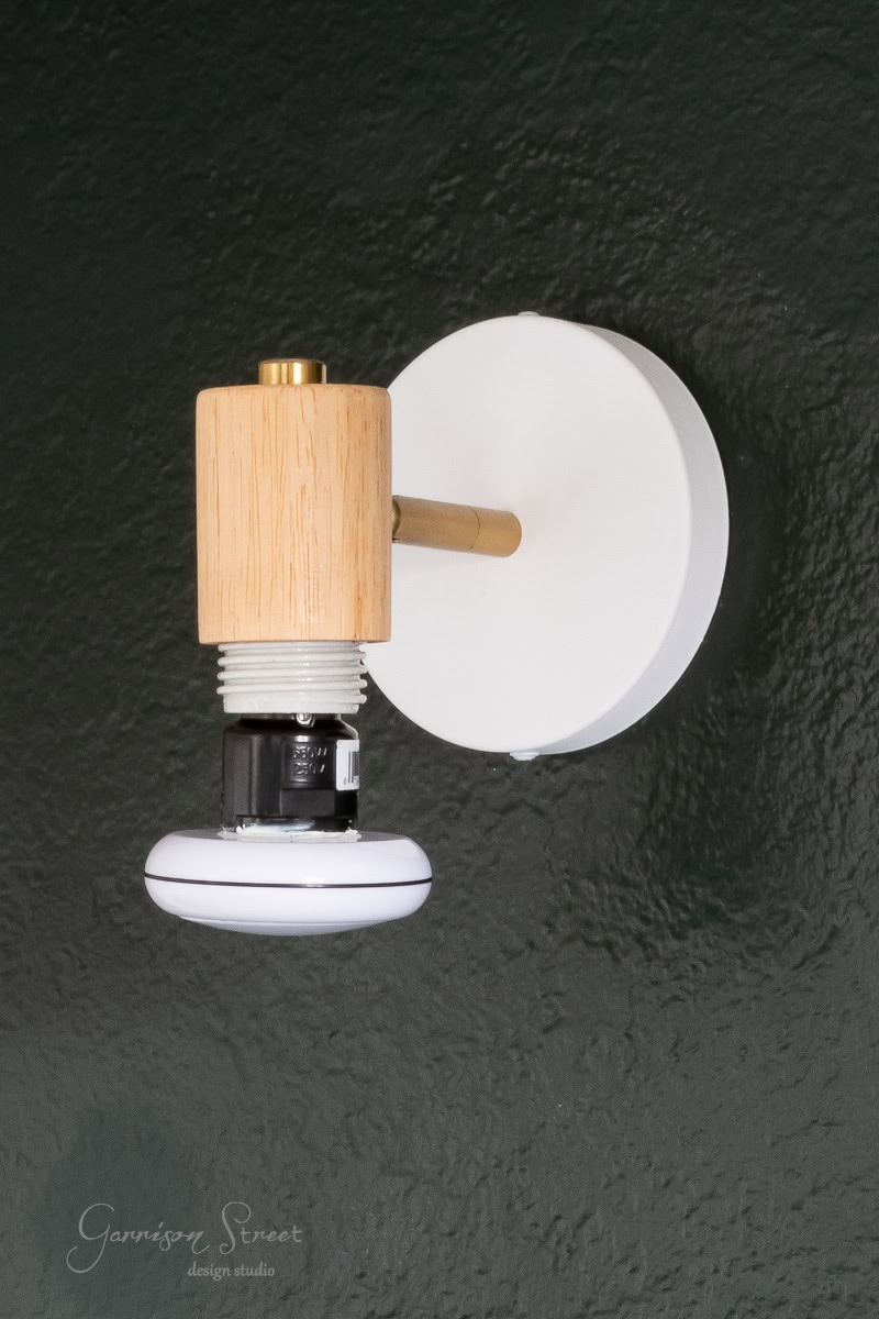 A modern DIY wall light fixture with a white circular base and a wooden cylindrical accent. This wireless sconce features a white, flat round bulb at the bottom, set against a dark green textured wall, blending functionality with style.