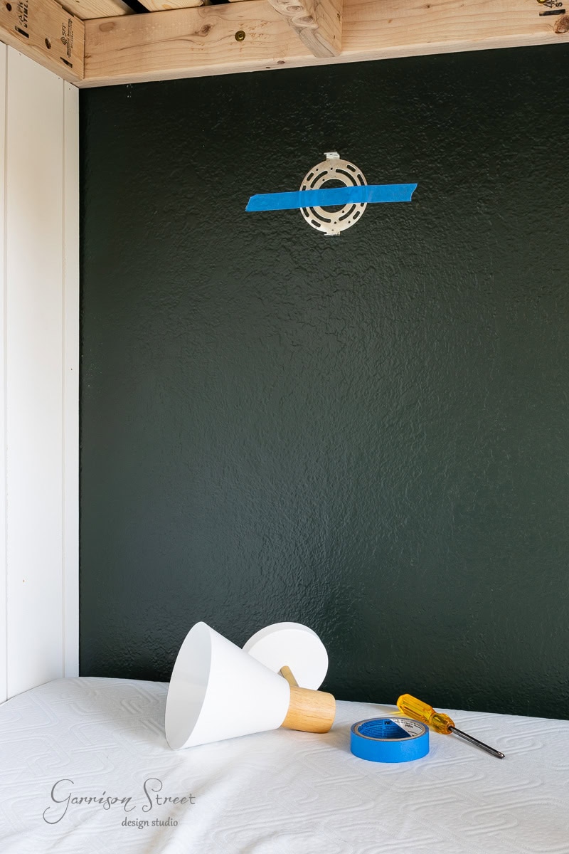 A partially assembled DIY wireless wall sconce rests on a covered surface against a dark green wall. A mounting bracket with blue tape is visible on the wall, while a screwdriver and a roll of blue tape sit nearby, ready to complete the installation.