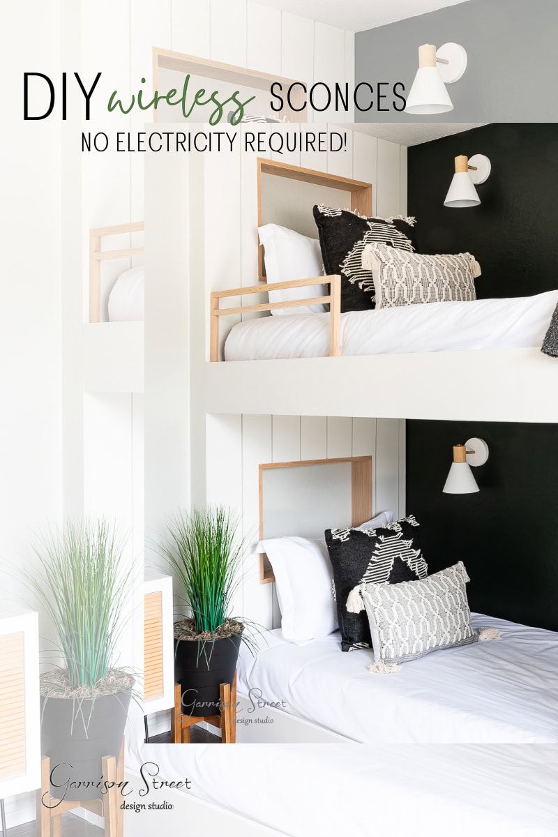 A modern bedroom showcases two white bunk beds adorned with black and white pillows and throws. Each bed has a DIY wireless wall sconce above, illuminating a green plant in a decorative pot. Text overlay reads: "DIY wireless sconces, no electricity required!."