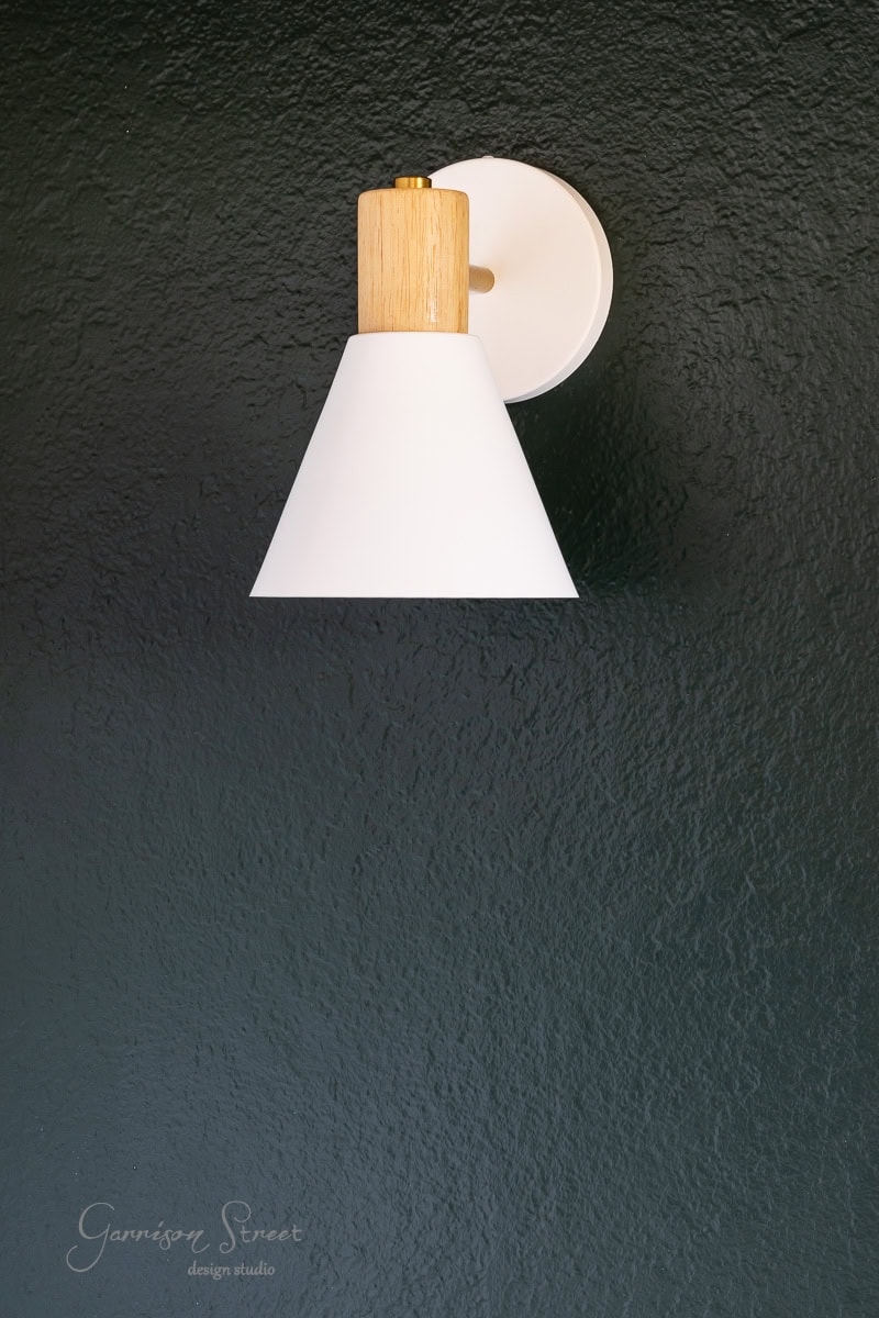 A modern wall light fixture, featuring a white conical shade and wooden accent, effortlessly enhances the textured dark green wall.