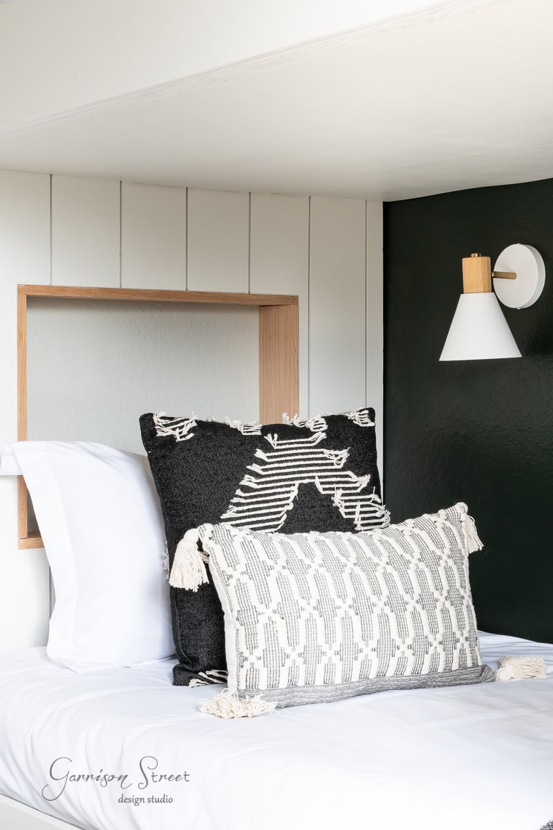 A cozy bed with white sheets and two decorative pillows—one black with a white geometric pattern and another white with a black pattern. A wooden wall alcove serves as a headboard, complemented by a DIY wireless wall sconce casting a gentle glow.