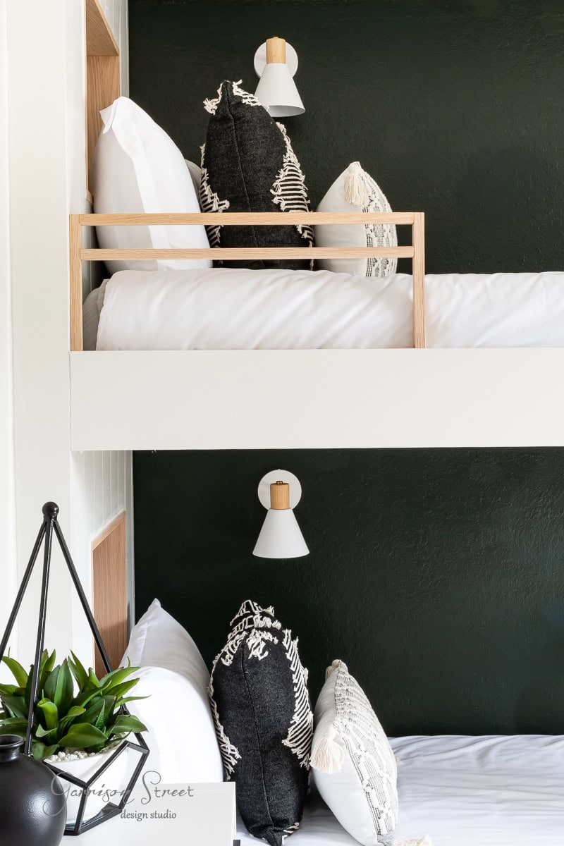 A cozy room features white bunk beds against a dark green wall, each adorned with patterned black and white pillows and a DIY wireless wall sconce above. A potted plant rests on the bottom bed, adding a touch of greenery.