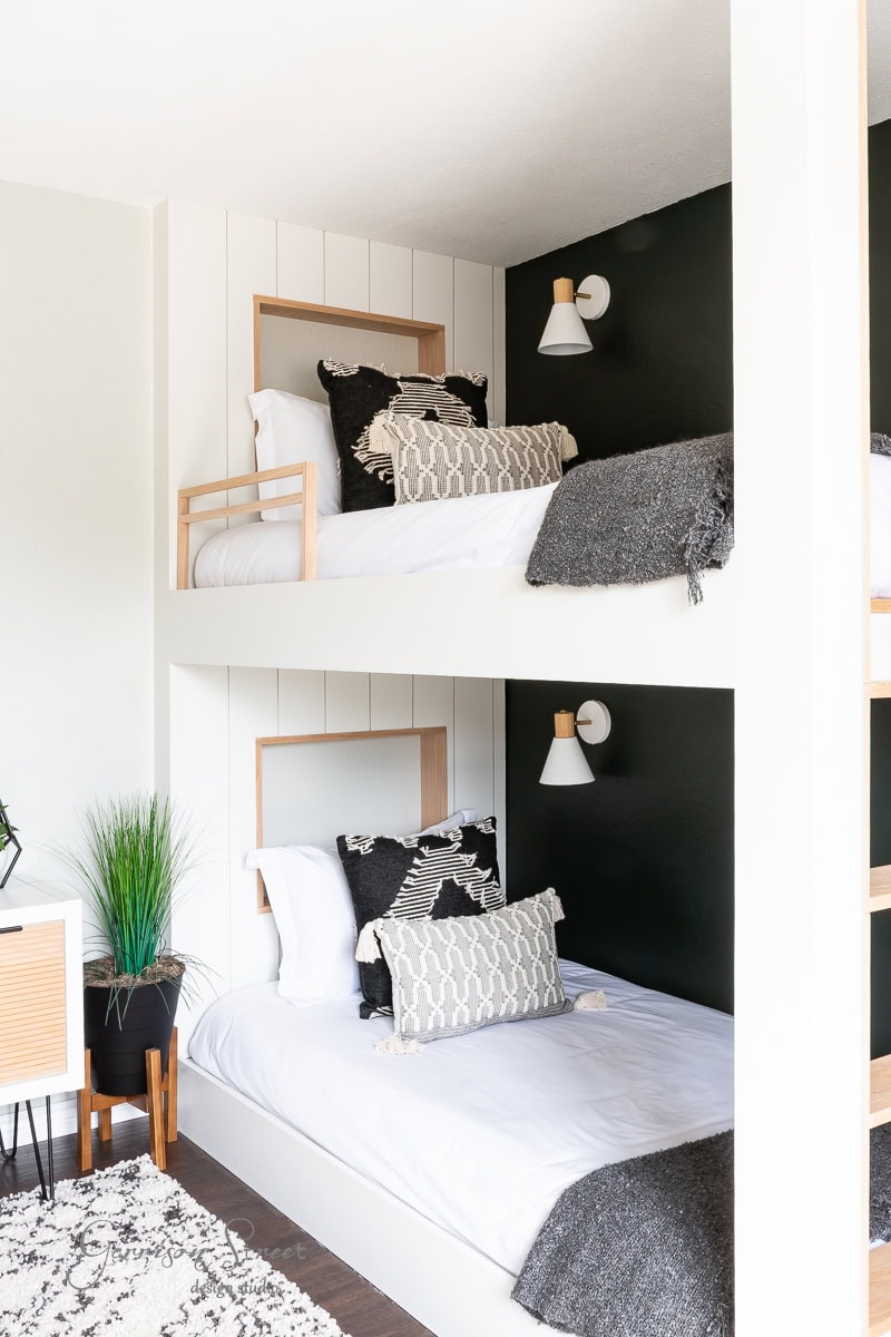 A modern bedroom with white walls features a bunk bed adorned with dark and light pillows and blankets. The room includes a wooden nightstand, a plant in a black pot, and stylish DIY wireless wall sconces above each bed. A beige and brown rug covers the floor.