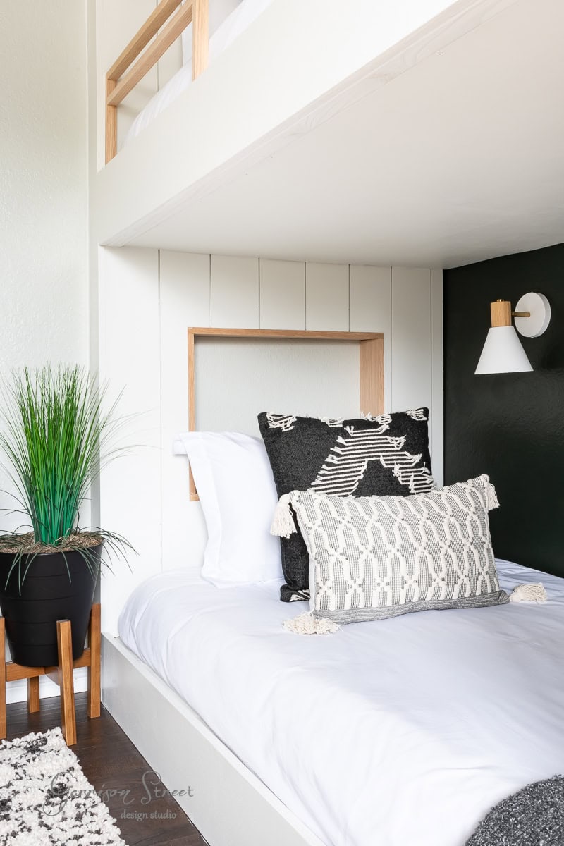 A modern bedroom features a white bed adorned with patterned pillows. A potted plant rests on a wooden stool beside the bed, while a DIY wireless wall sconce lights up the dark accent wall. Above, a loft with a wooden railing adds character to the space.