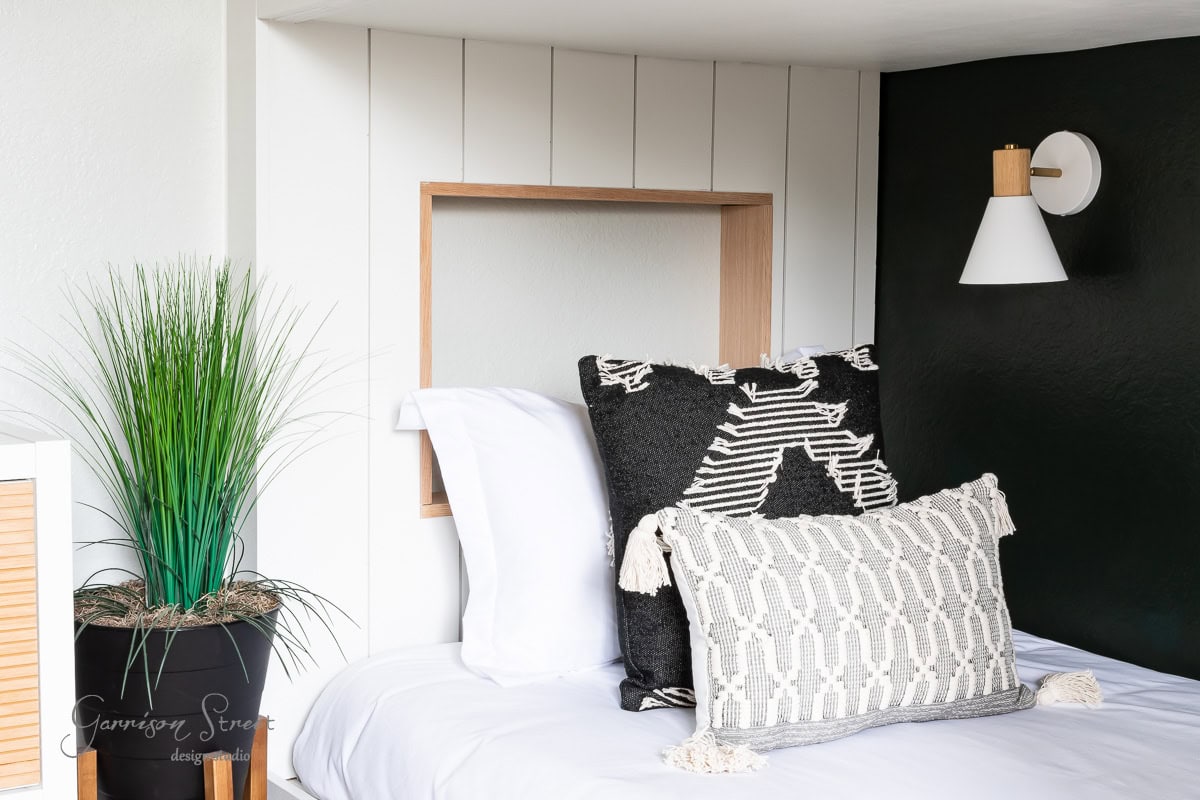 A modern bedroom features a black accent wall adorned with a DIY wireless wall sconce. A stylish bed is decked out with white and black geometric-patterned pillows, while a potted plant with green and blue grasses adds a touch of nature beside it.