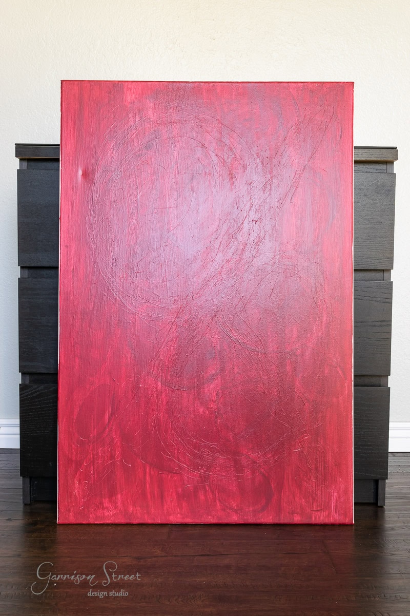 A large abstract painting featuring swirling red brush strokes on a canvas propped against a dark wooden cabinet. The vibrant red artwork contrasts with the neutral background and dark furniture, creating an effect reminiscent of DIY fabric wall art.