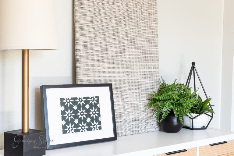 A modern interior setup features a lamp, a black and white geometric framed print, DIY fabric wall art with a textured pattern, and two potted plants on a white shelf. The arrangement is minimalistic and aesthetically pleasing.