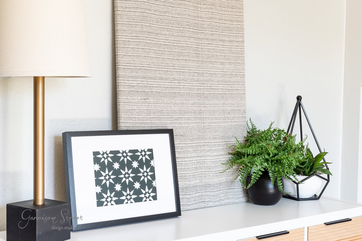 A modern interior setup features a lamp, a black and white geometric framed print, DIY fabric wall art with a textured pattern, and two potted plants on a white shelf. The arrangement is minimalistic and aesthetically pleasing.