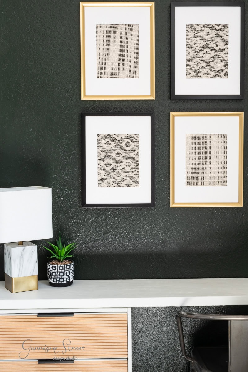 A modern interior with a dark green wall showcases DIY fabric wall art in gold and black frames. Below, a white desk holds a marble lamp, a small plant, and a black chair. The desk's drawers feature a wood texture.