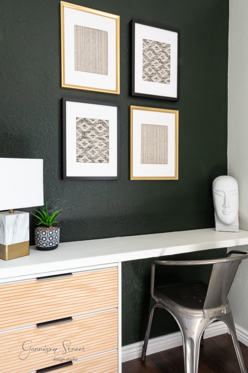 A modern office corner features a white desk with a sleek metal chair. Above the desk, four framed abstract artworks include DIY fabric wall art, adding a personal touch. A sculptural white lamp and a small plant decorate the desk. The dark green wall complements the minimalist vibe perfectly.