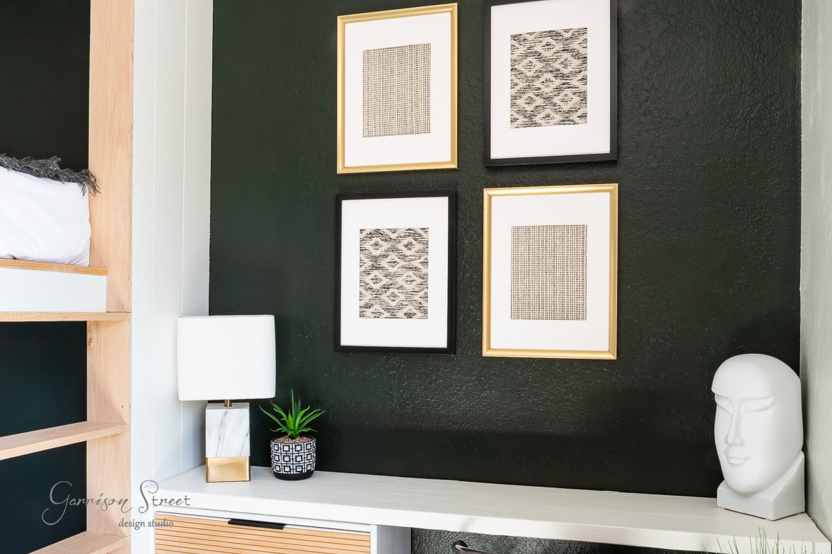 A modern interior showcases a dark green wall adorned with DIY fabric wall art arranged in a grid. Below, a sleek white desk hosts a matching lamp, a small green potted plant, and a decorative white sculpture. A hint of a bunk bed peeks from the left.