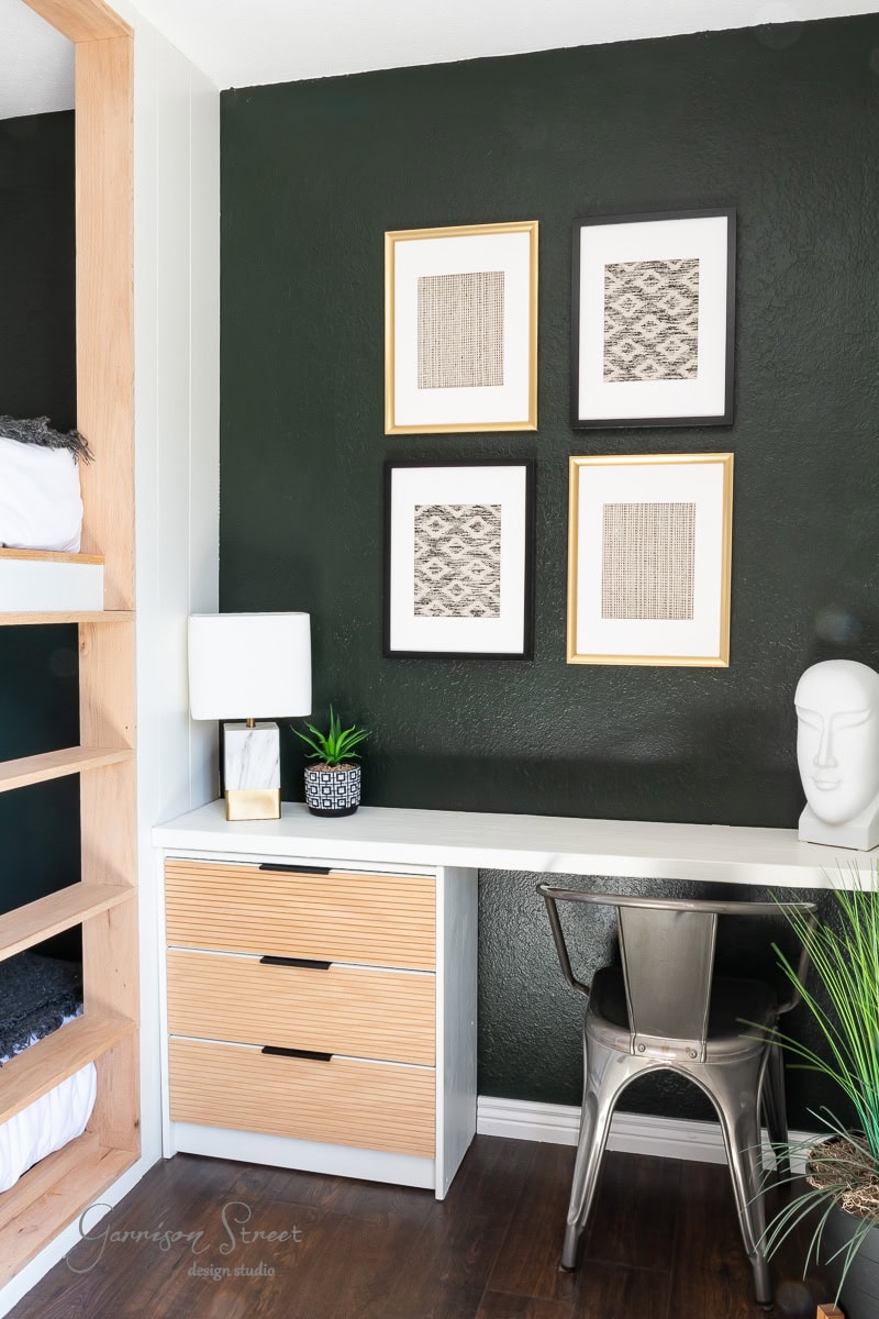 Minimalist workspace with a white desk, metal chair, and a wooden drawer unit. A lamp, small plant, and statue sit on the desk. Four framed abstract artworks adorn a dark green wall, including DIY fabric wall art. A built-in wooden feature with bedding is adjacent.