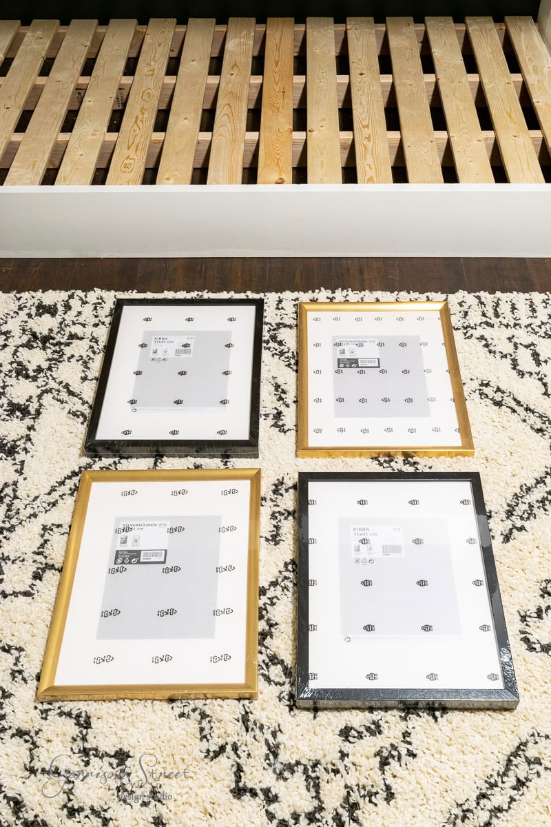 Four picture frames displayed on a patterned black and white rug bring an artistic touch to DIY fabric wall art. Two frames are gold, two are black, and behind them stands a wooden bed frame without a mattress, awaiting its cozy finishing touches.