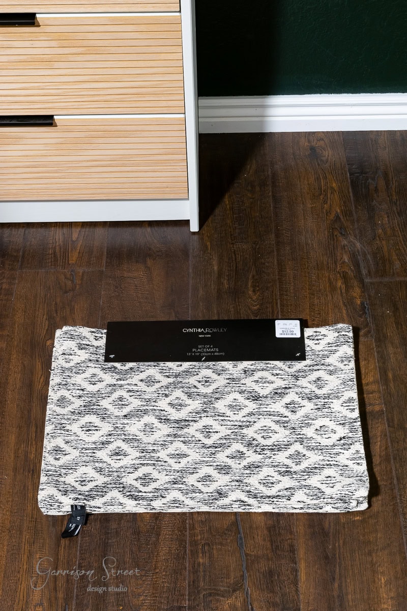 A black and white patterned rug is placed on a dark wooden floor, partially unrolled with a label attached. Nearby, a light-colored wooden cabinet with drawers offers the perfect spot to showcase DIY fabric wall art.