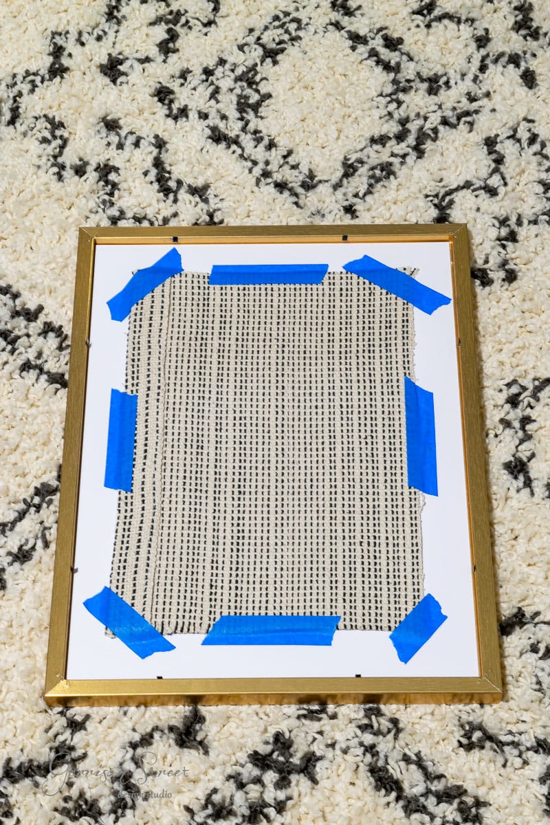 A DIY fabric wall art featuring a black dotted pattern is framed with a gold edge. It's fixed to a white board using blue tape and placed on a textured, beige and black patterned carpet.