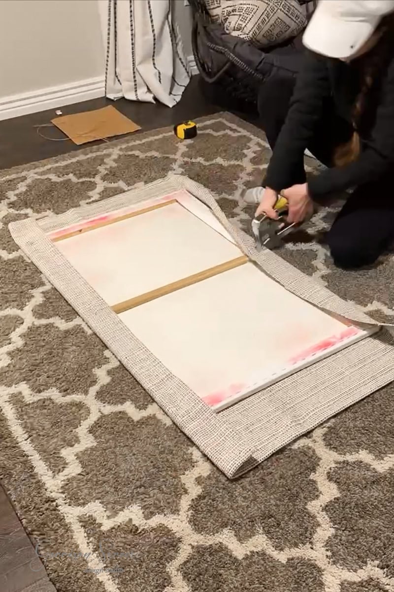 A person is upholstering a rectangular cushion with beige fabric on a patterned rug, creating the perfect accent for their DIY fabric wall art. They use a staple gun to secure the material to the cushion's backside, with a measuring tape and cardboard piece visible nearby.