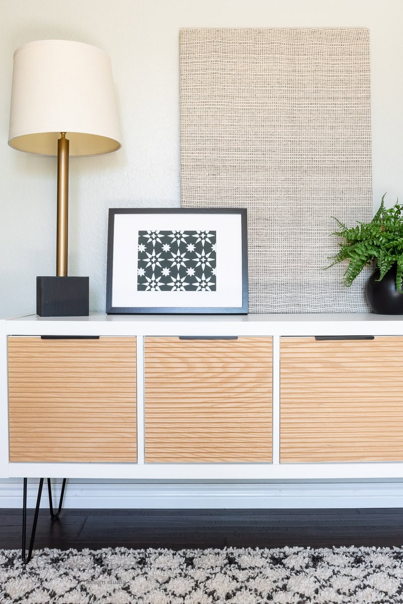 A modern, minimalist room featuring a wooden console with geometric-patterned artwork, a bronze lamp with a white shade, and a green plant. Above hangs DIY fabric wall art in neutral tones, complementing the patterned rug below.