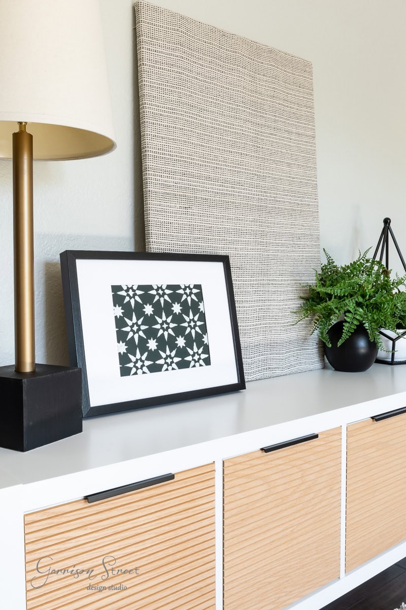 A modern interior scene showcases a white console table adorned with a geometric black and white framed artwork, a lamp with a black base, and a lush green plant in a black pot. A DIY fabric wall art panel adds texture as it leans gracefully against the wall.