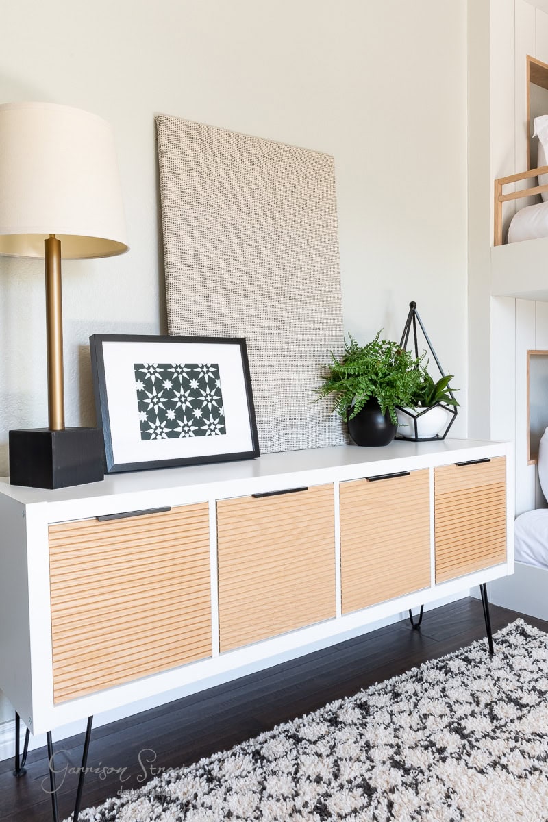 A modern room showcases a white sideboard with wood-paneled doors on slim legs. It elegantly displays DIY fabric wall art, a framed geometric print, and a potted fern. A black and gold lamp completes the look, while the floor is adorned with a patterned rug.