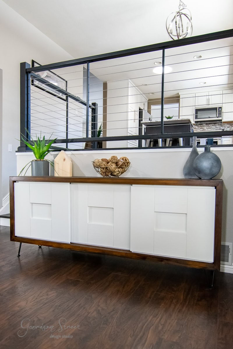 Modern interior with a mid-century style sideboard featuring white paneled doors and dark wood frame, exemplifying cost-effective decor solutions. Decorative items, including a potted plant and bowls, sit on top. Behind, a kitchen area is visible through a wire and metal partition.