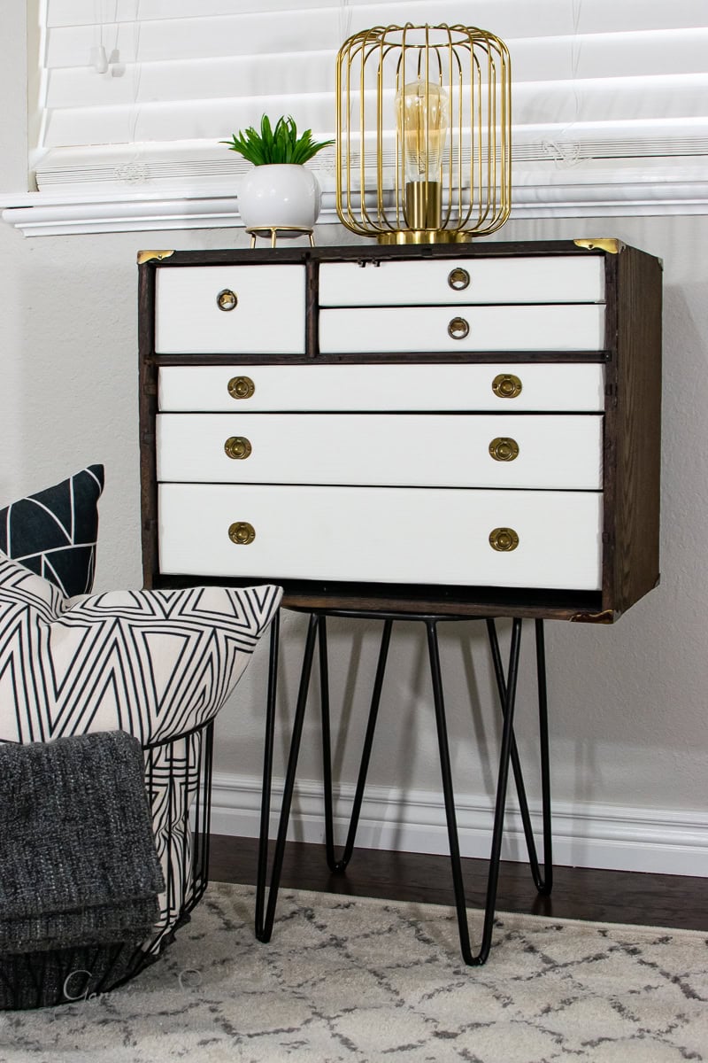 A small cabinet with white drawers and gold knobs stands on hairpin legs, offering a glimpse of style for those wondering how much to furnish a 4-bedroom house. It holds a potted plant and a bulb encased in metallic wire, while nearby, a patterned cushion and folded blanket rest on a textured rug.