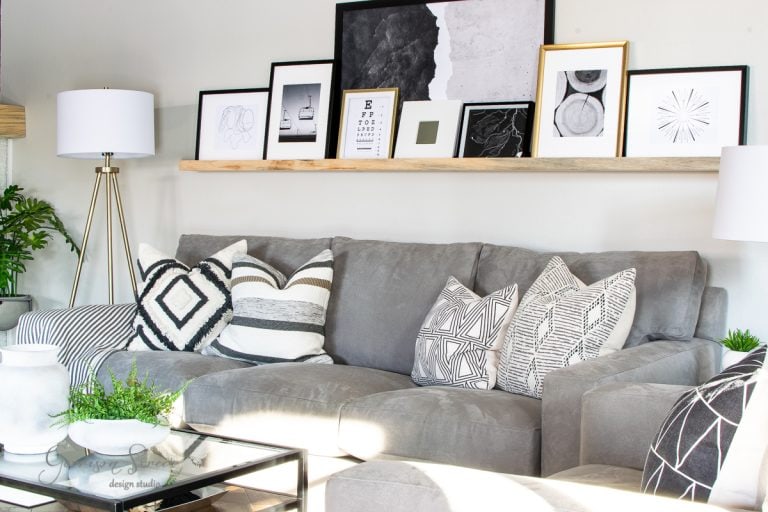 A modern living room features a gray couch with patterned pillows. Above, a wooden shelf showcases framed abstract art and photographs. A floor lamp with a white shade stands nearby, while a glass coffee table sits in front, capturing the essence of how much to furnish a 4-bedroom house.