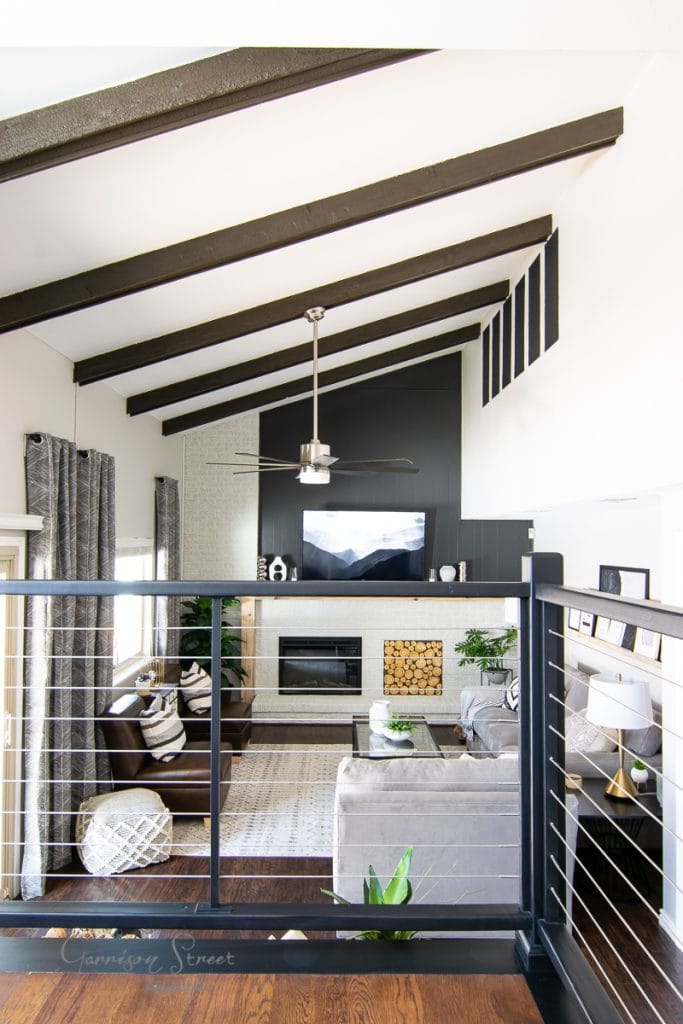 A modern living room with a vaulted ceiling and exposed beams showcases a large gray sectional sofa, an elegant fireplace against a decorative black wall, and a sleek ceiling fan. Natural light floods through expansive windows, while lush greenery injects a trace of nature—consider these elements when budgeting to furnish a 4-bedroom house.